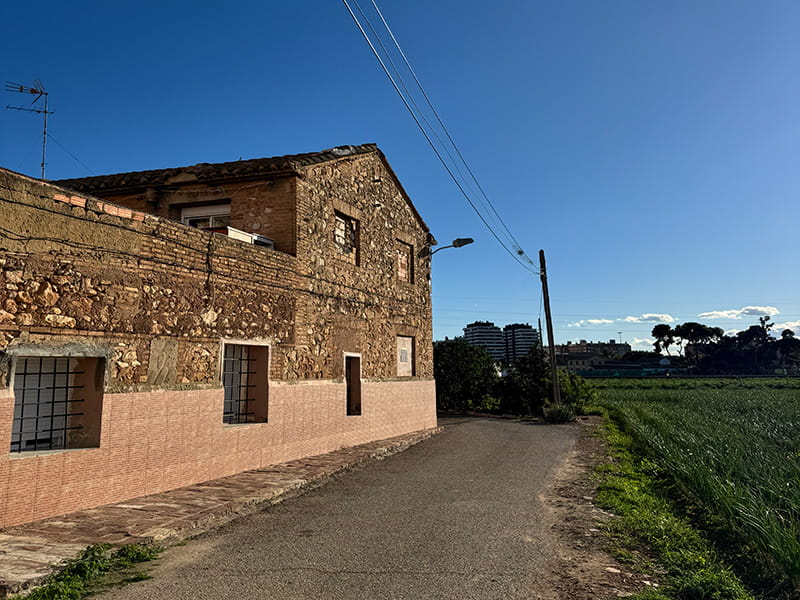 Старая каменная алькерия у узкой сельской дороги среди полей уэрты Pobles del Nord; вдали виден городской горизонт Валенсии и линии электропередачи на фоне ясного неба.